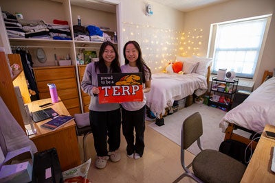 Two smiling female students in a residence hall room holding a "Proud to be a TERP" sign.