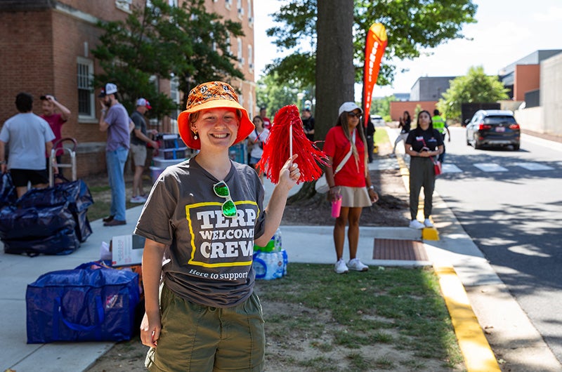 A smiling volunteer in a Terp Welcome Crew shirt and orange bucket hat holds a red pom-pom.