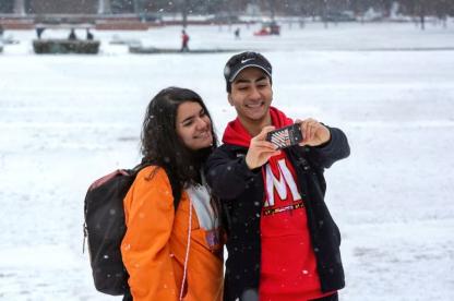 two students take a selfie as snow falls overlooking the Mall with Main Administration Building in the background.