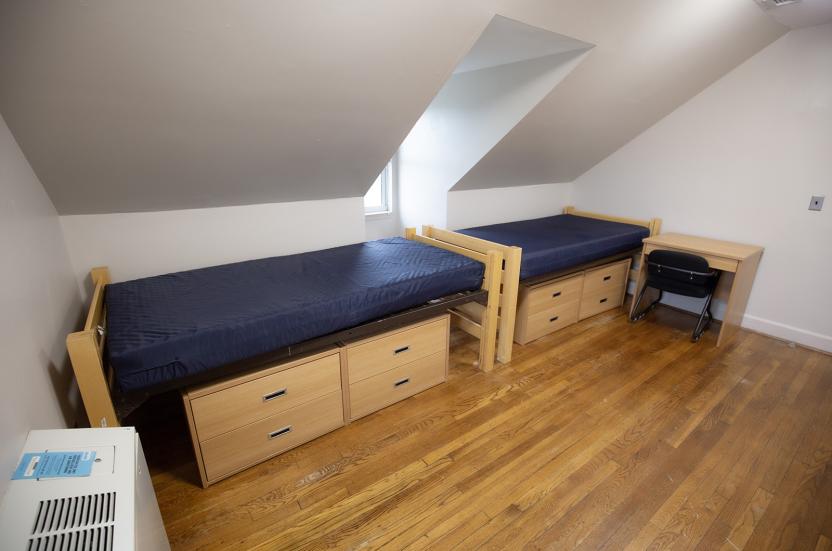 Double residence hall room with angled ceiling, two twin beds with drawers underneath, and a desk.