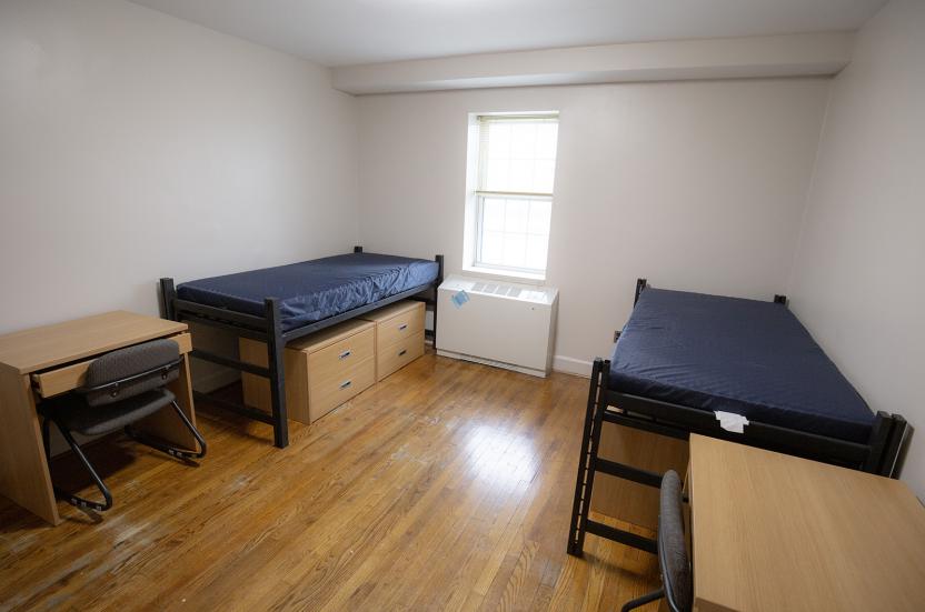 Double residence hall room with hardwood floors, two twin beds with small dressers underneath, and two study desks with chairs.