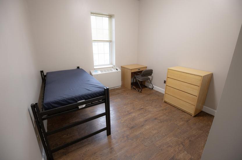Single residence room with wood flooring, a window, a twin bed, a desk with a chair, and five-drawer dresser.