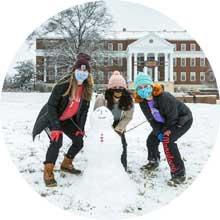 Three female students wearing masks building a snowman on the Mall; McKeldin Library in the background.