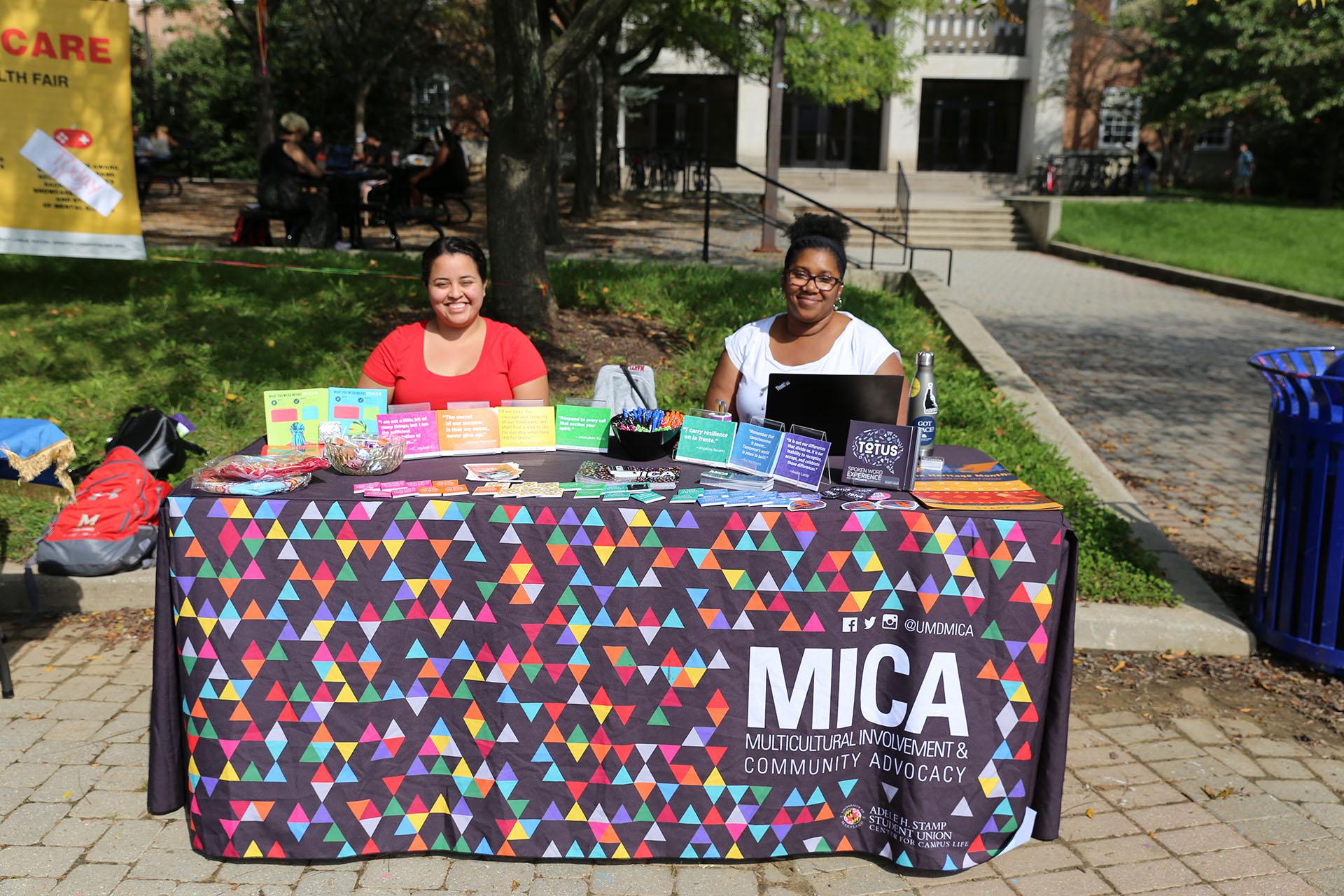 two people smiling behind an foldout table outside and draped with a cloth that reads MICA