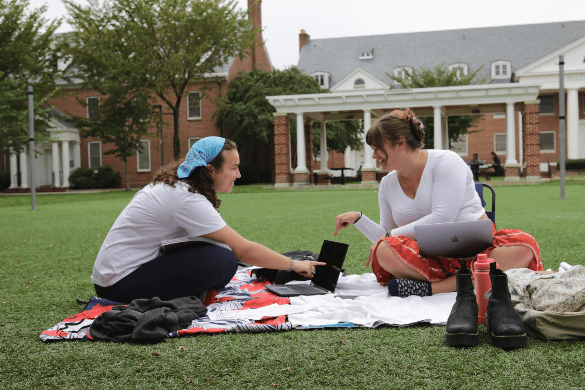 two students sitting on a blanket on the grass at Washington Quad smiling and pointing to laptop