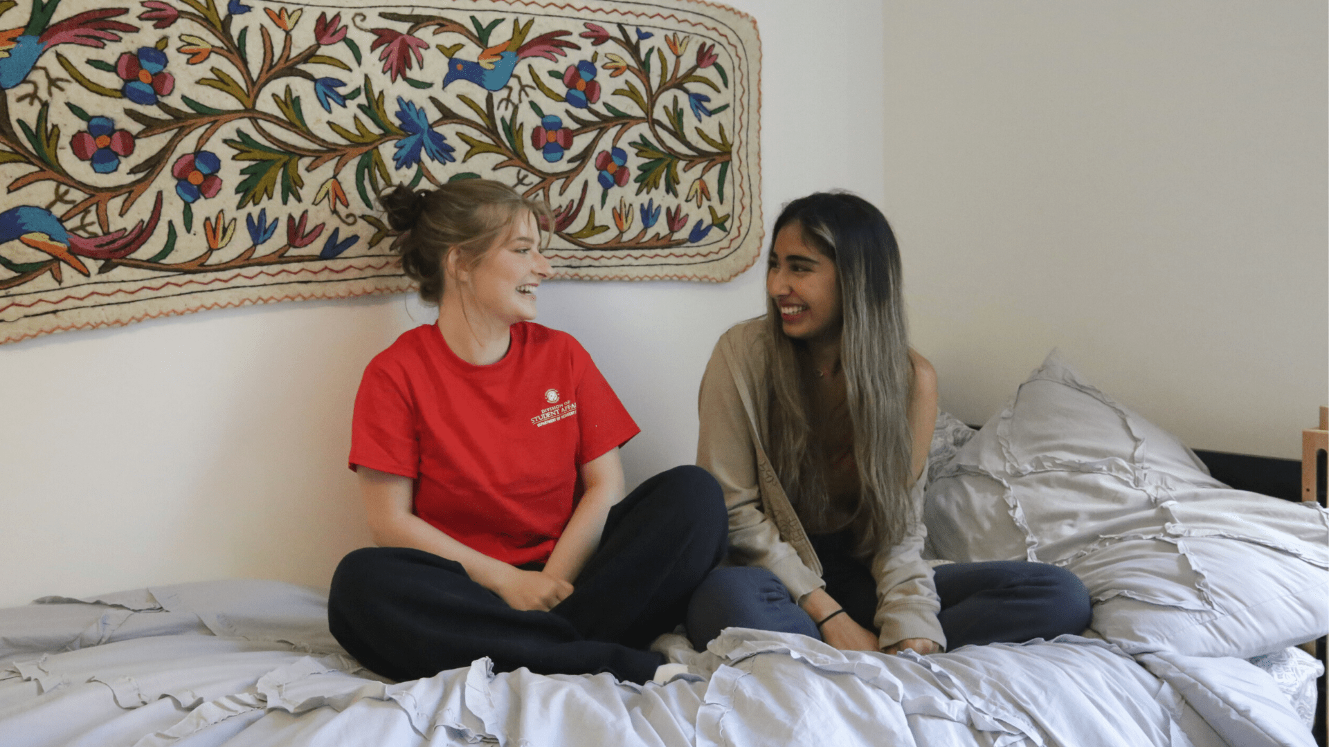 two students in dorm room chatting on a bed