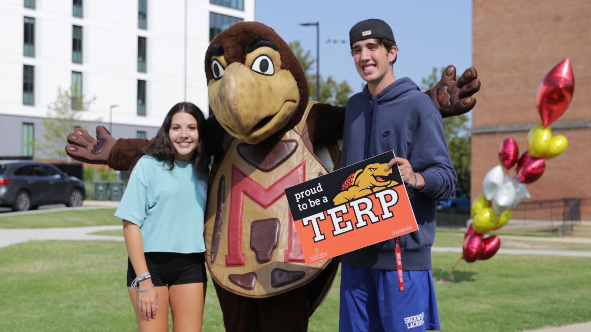 testudo mascot with two students outside on move-in day, balloons and residence halls in the background
