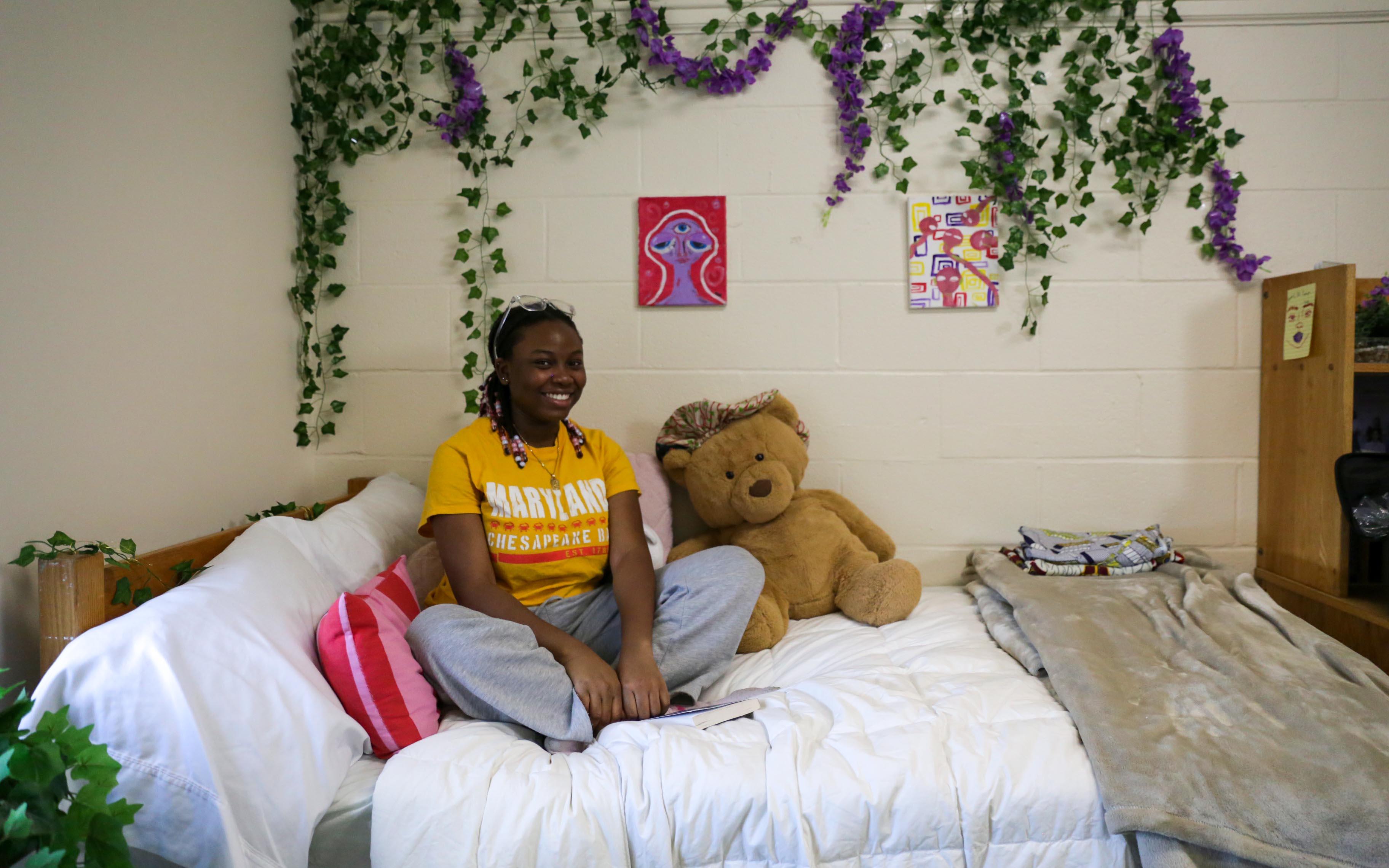student sitting on her bed decorated with blanket and teddy bear in residence hall room, hanging wall plants in the background 
