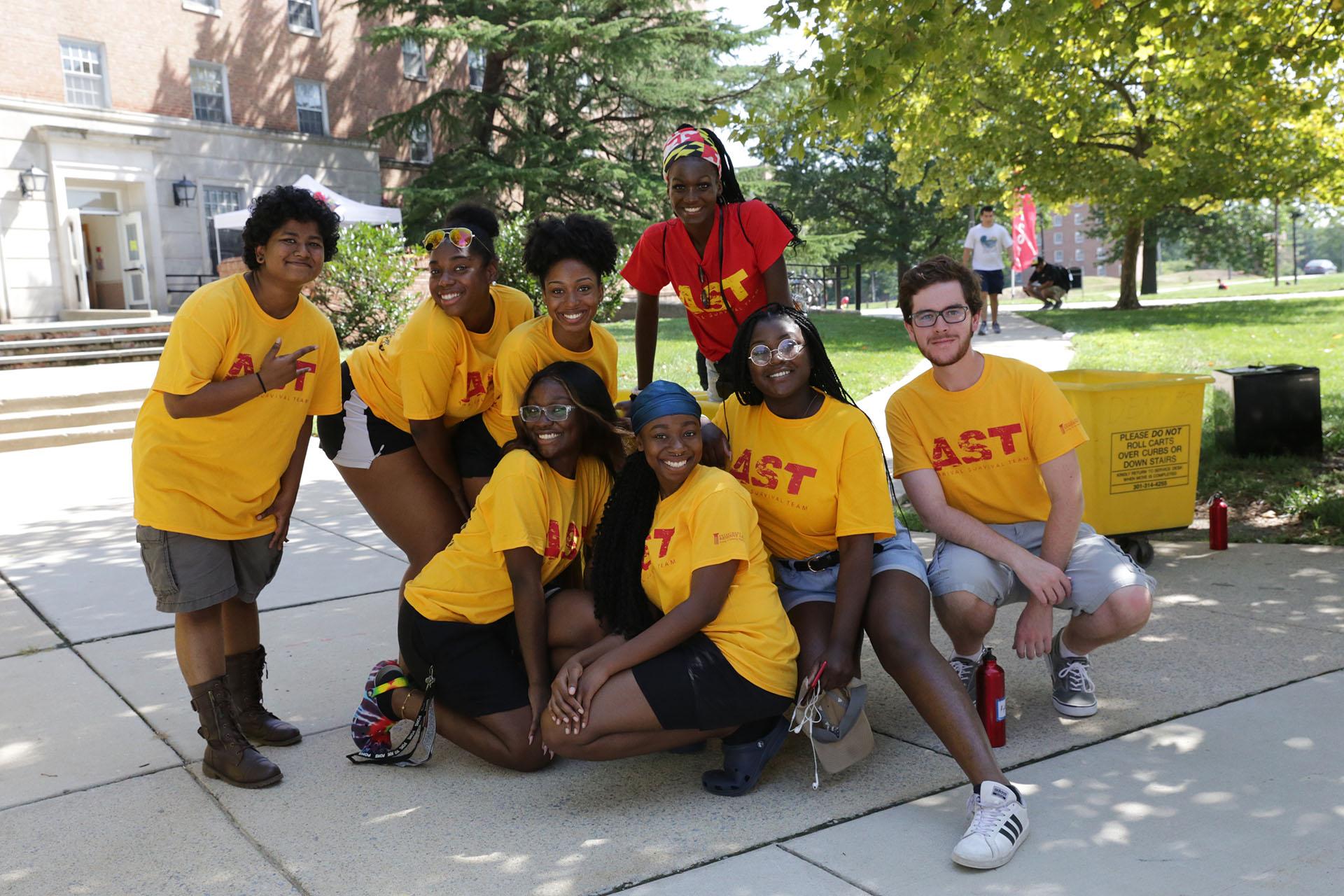 diverse group of eight students posing outside residence hall during fall move-in, seven are wearing shorts and yellow shirt and another student is wearing a red shirt