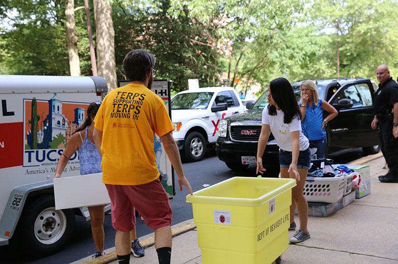 student with back facing camera wearing a yellow shirt and pushing a cart on curbside while other people load belonging out of car