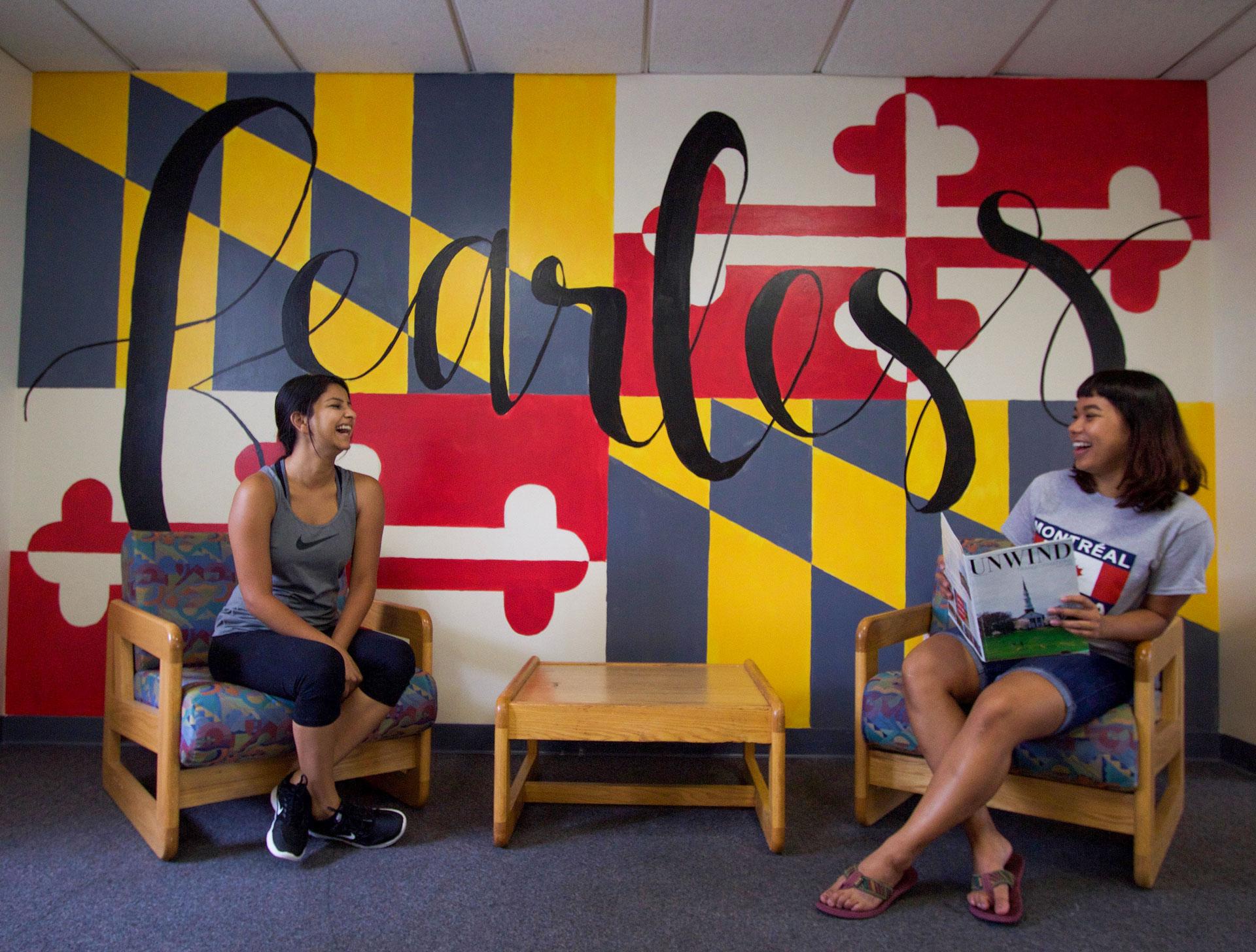 two students sharing a laugh sitting side-by-side on chairs in front of a wall decorated with the Maryland flag and word fearless