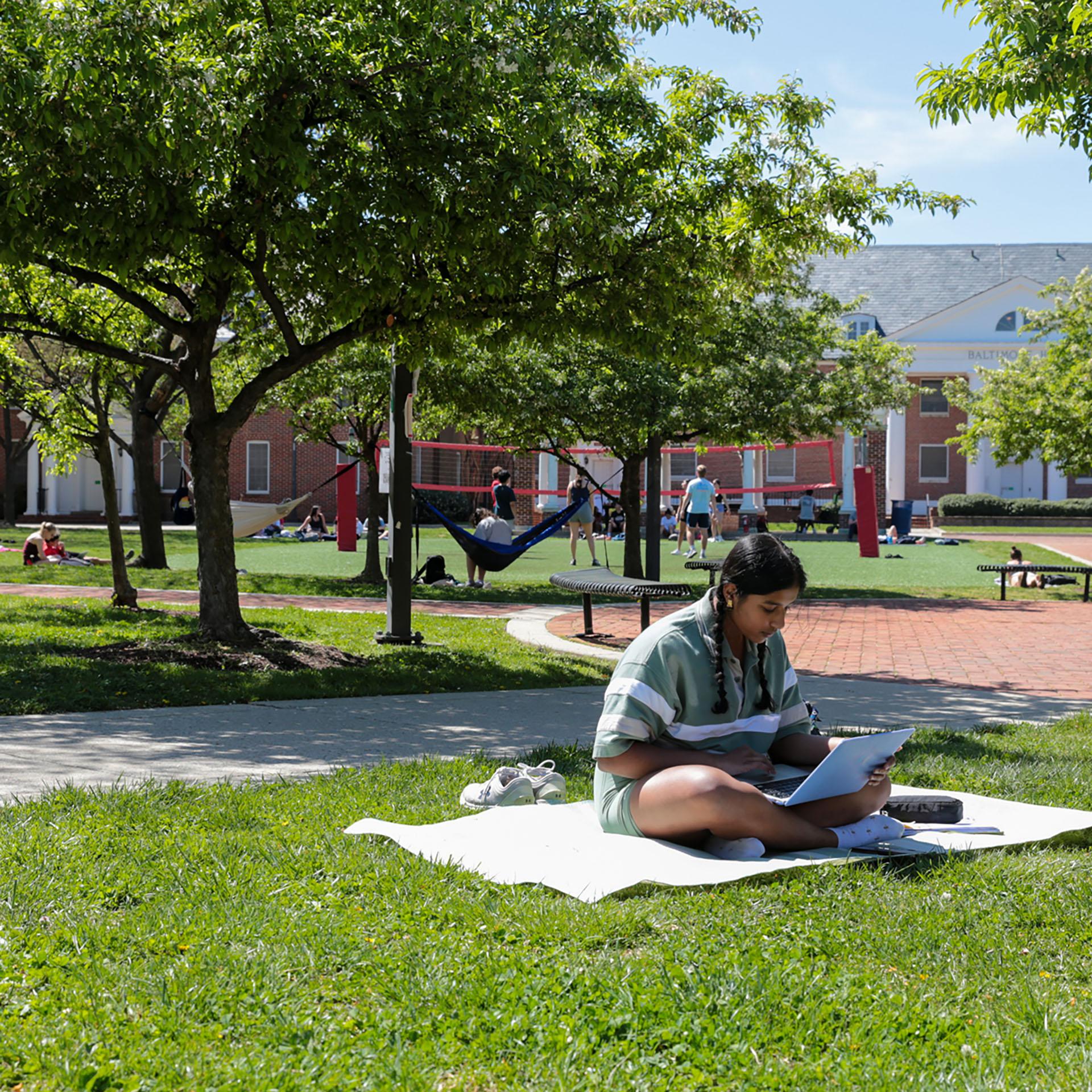 Student sitting on blanket using laptop in Washington Quad in the foreground with students playing volleyball and using hammocks in the background