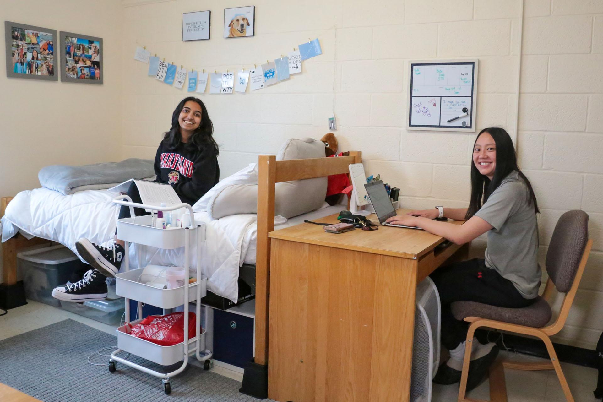 two students in residence hall room, one sitting on a bed and another at the study desk