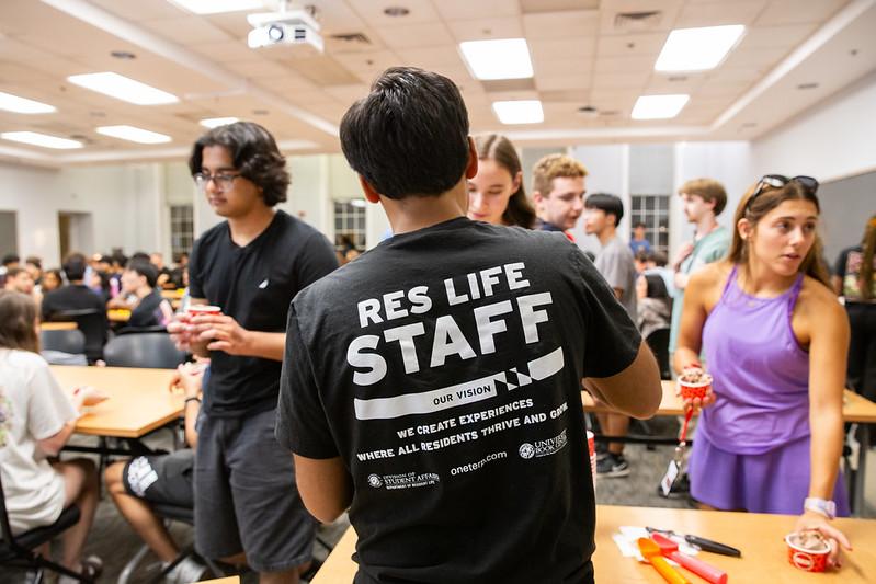 Student wearing a "RES LIFE STAFF" t-shirt in a room with other students.
