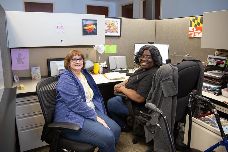 Two professional staff members smiling in an office cubicle.