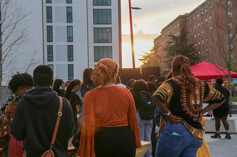 Students gathered outdoors at sunset, standing near benches and talking in front of campus residence halls.
