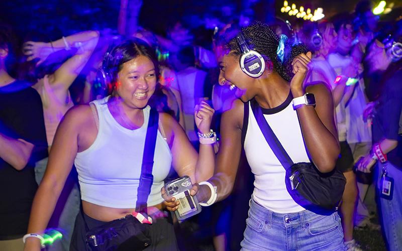 Two students wearing light-up headphones dance and laugh at an outdoor silent disco event.