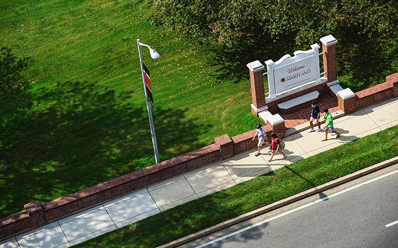 Aerial view of students walking on a campus sidewalk past a "Welcome to Maryland" sign.