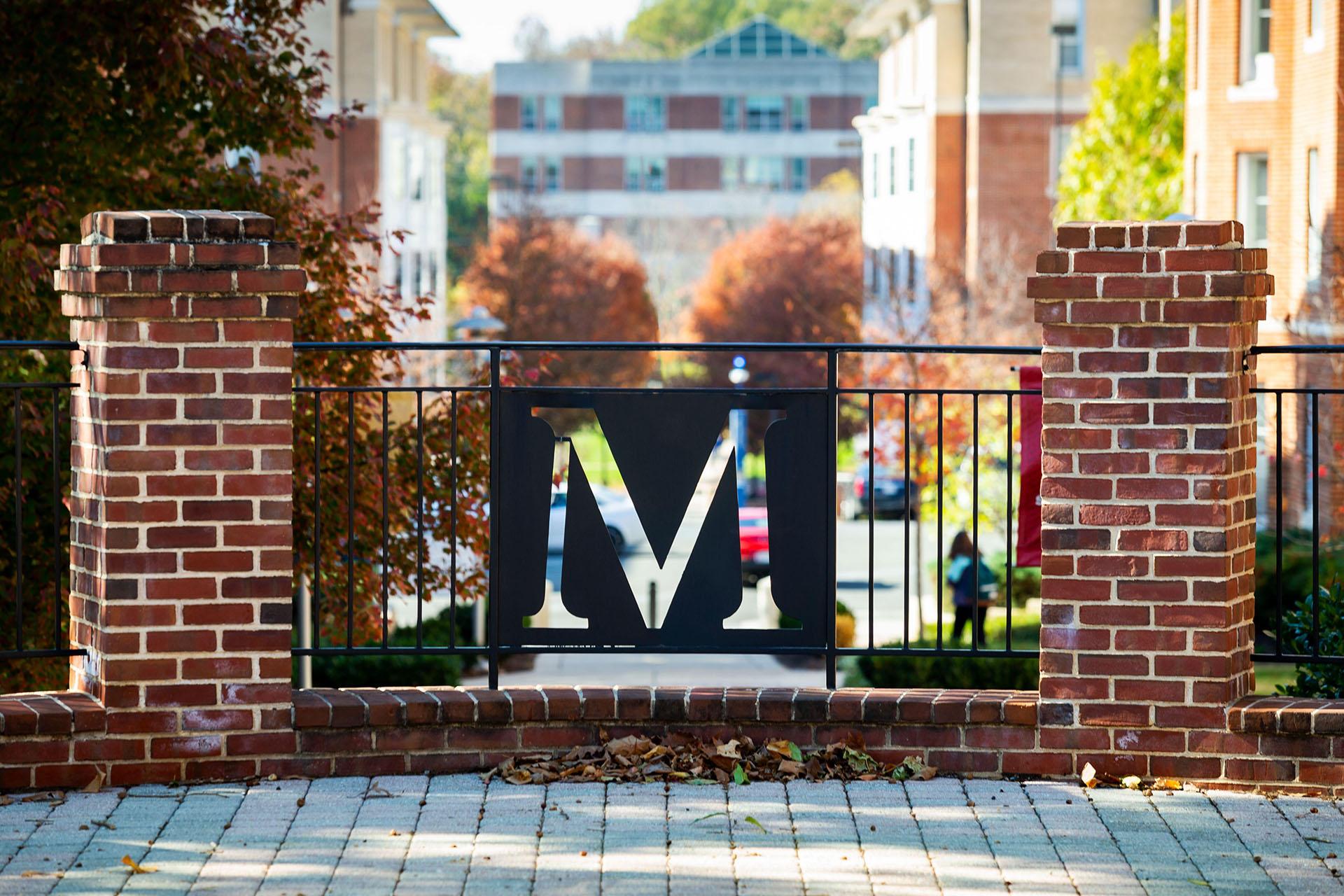 A black metal "M" logo set into a brick railing overlooks the South Campus Commons area.