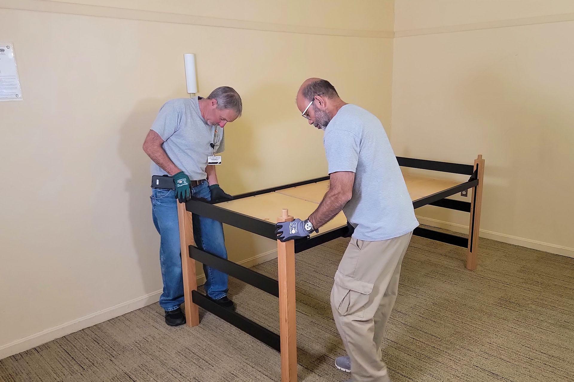 Two staff members in grey shirts and work gloves assemble a wooden loft bed frame in a room.