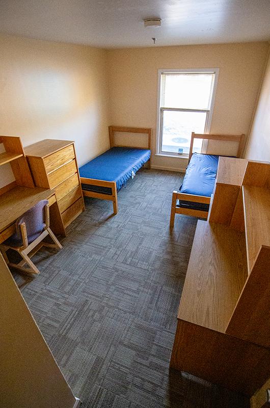 An empty double room with two blue-covered beds, wooden desks, and a window.