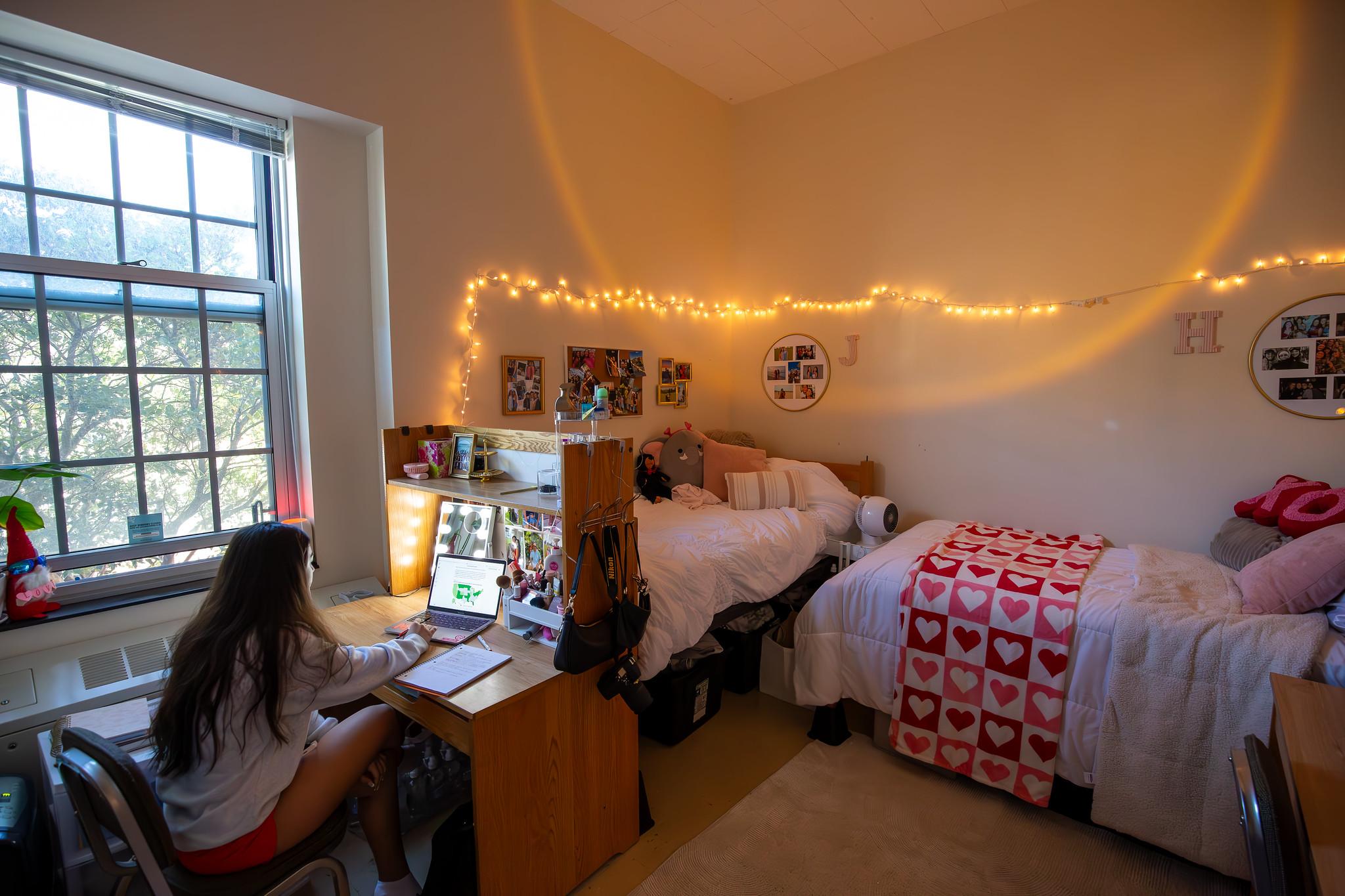 A student sits at a desk in a decorated residence hall room with string lights and heart-patterned bedding.