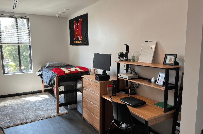 A tidy dorm room with a desk, a "M" flag on the wall, and a bed with a Maryland-themed blanket.