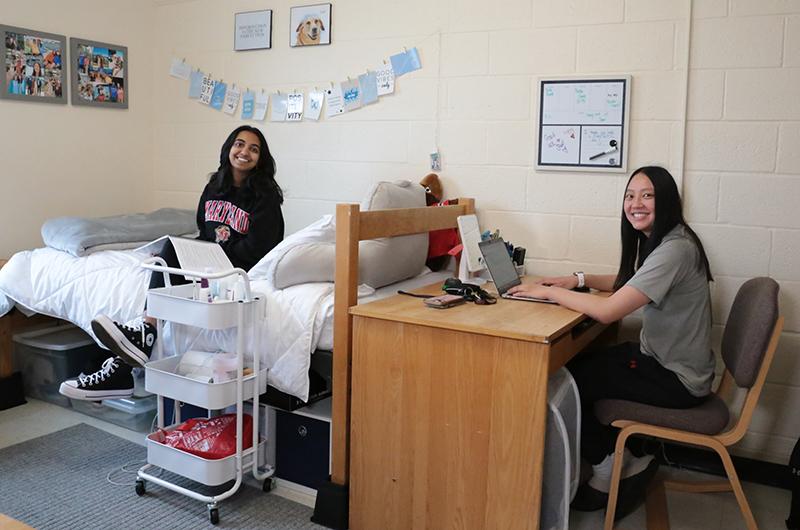Two smiling roommates in a decorated residence hall room, one sitting on a bed and one at a desk.