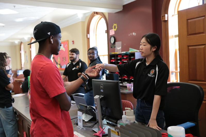 A community assistant in a black polo shirt hands room keys to a student at a residence hall service desk.