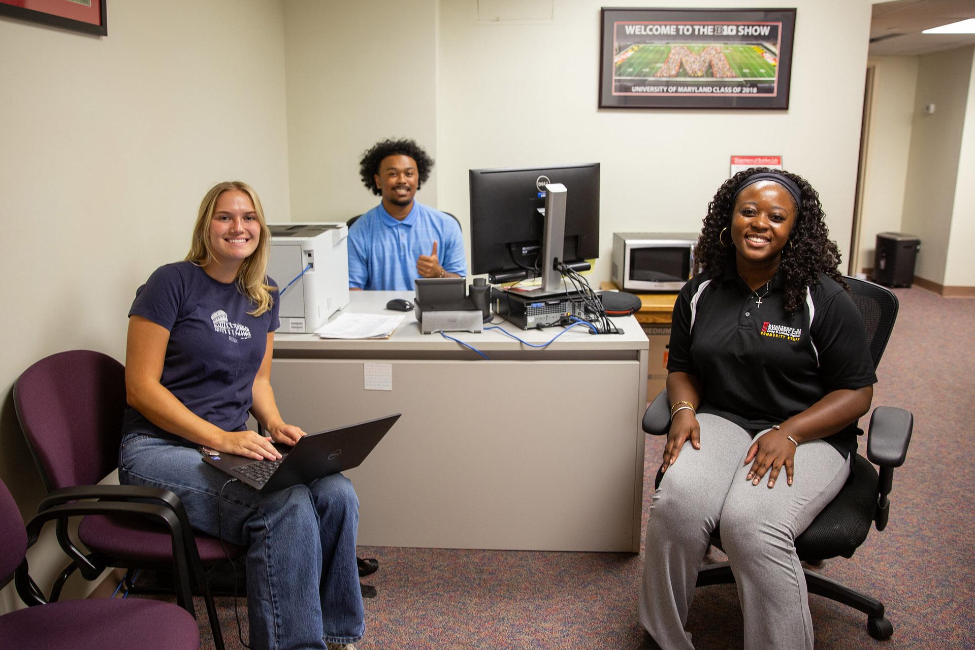 Three student staff members smile and pose near a desk in a university office setting.