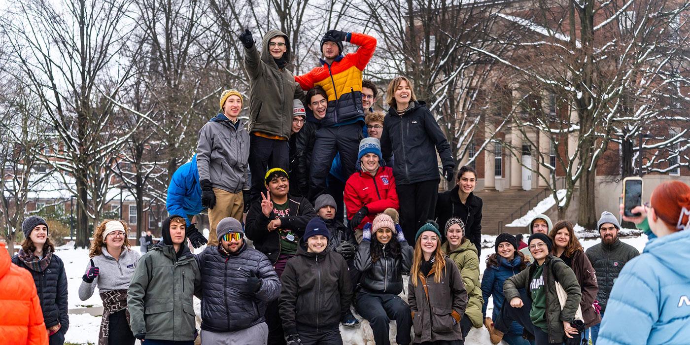 A large group of students in winter gear pose together on a snowy campus mall.