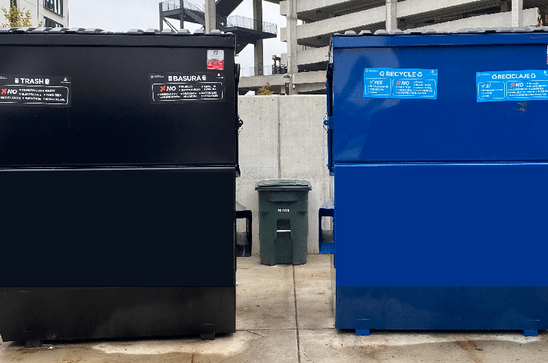 Large black trash and blue recycling dumpsters side-by-side with instruction labels in English and Spanish.
