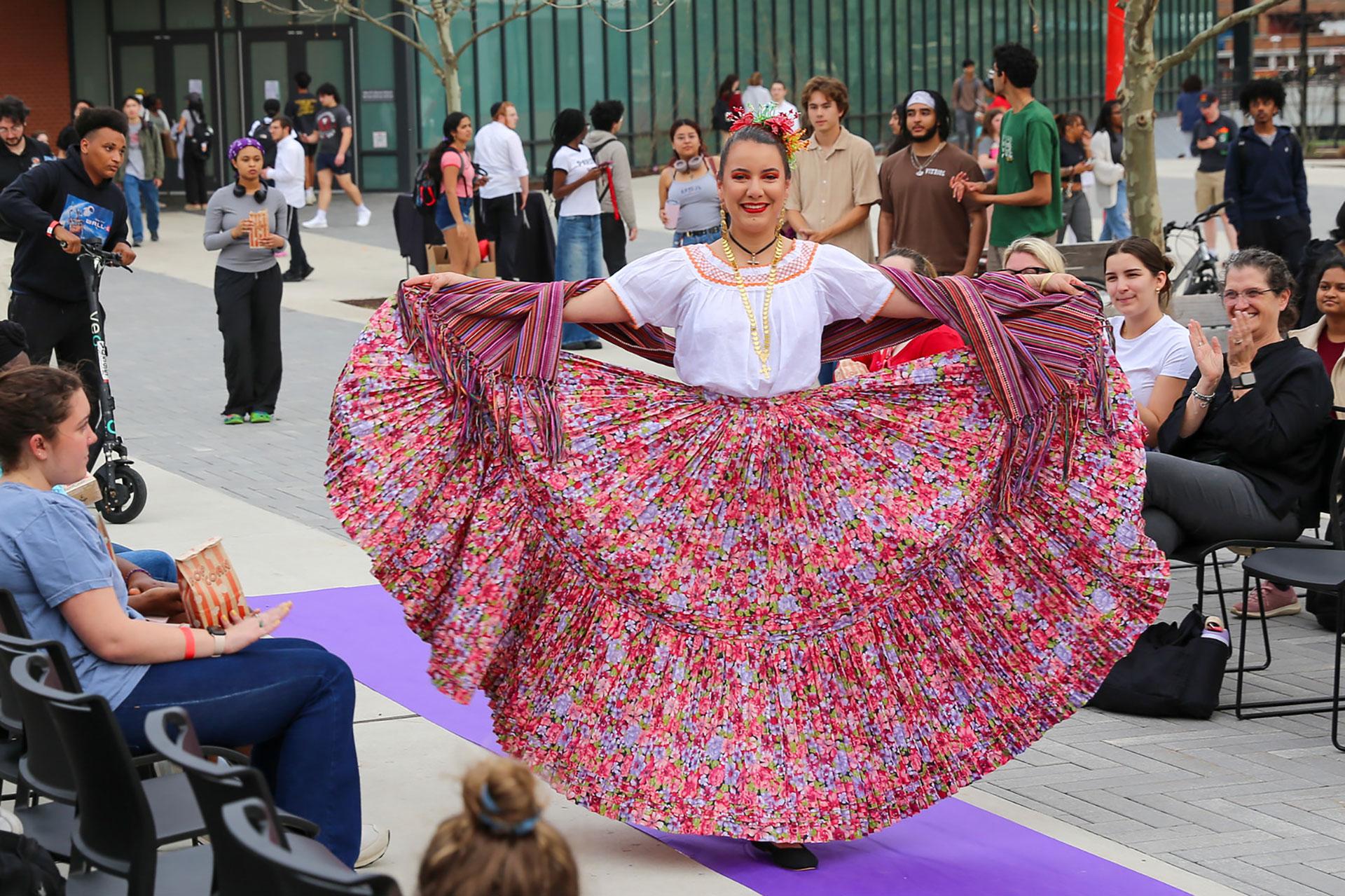 A smiling student in a vibrant, floral folkloric dress performs down a runway during Embrace the Heritage Showcase event.