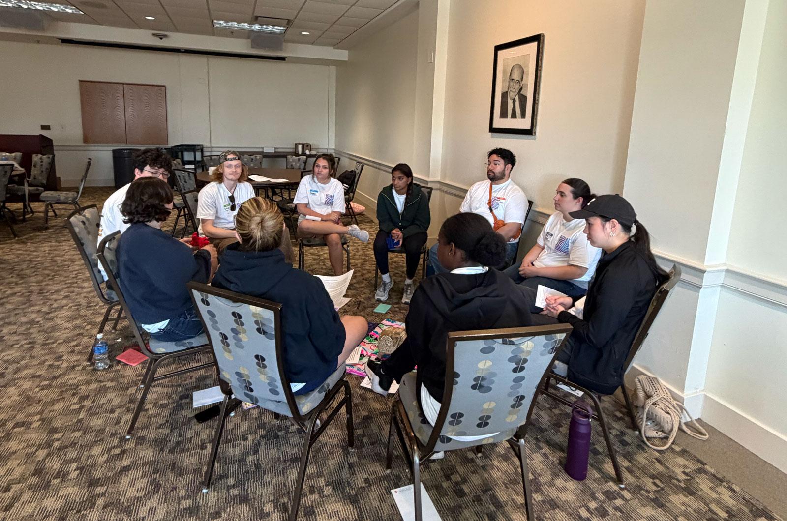 A group of volunteers sit in a circle of chairs for a restorative discussion in a carpeted meeting room.
