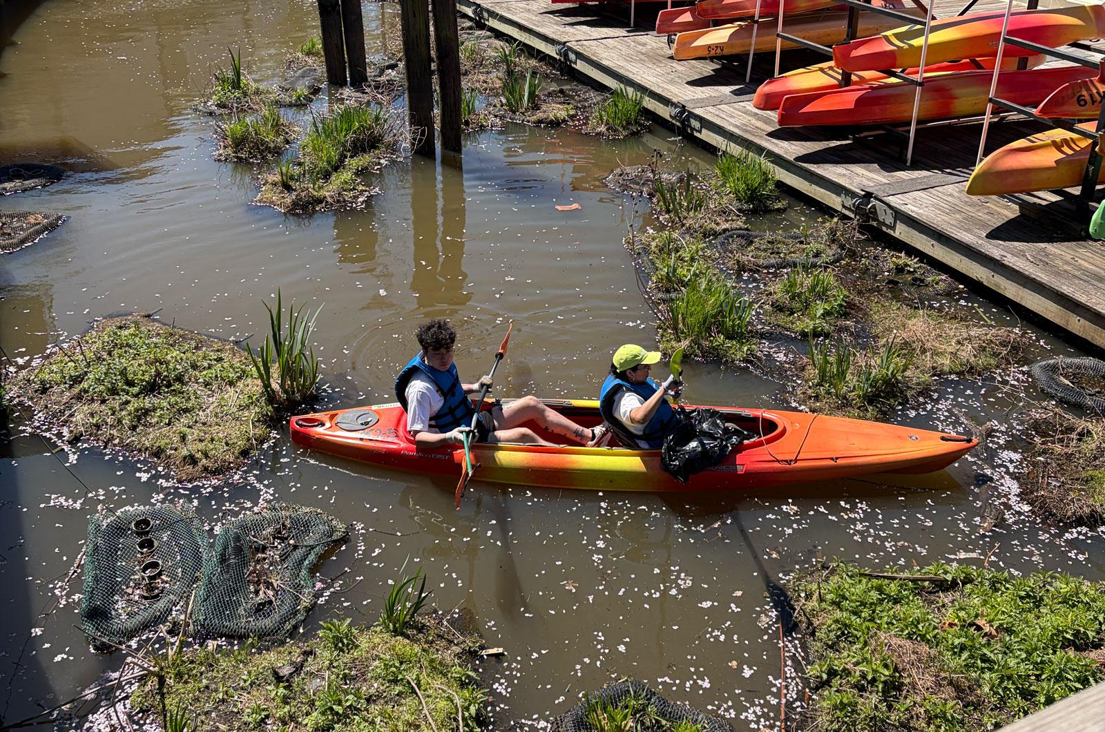 Two people in a tandem kayak paddle near a dock filled with colorful kayaks.