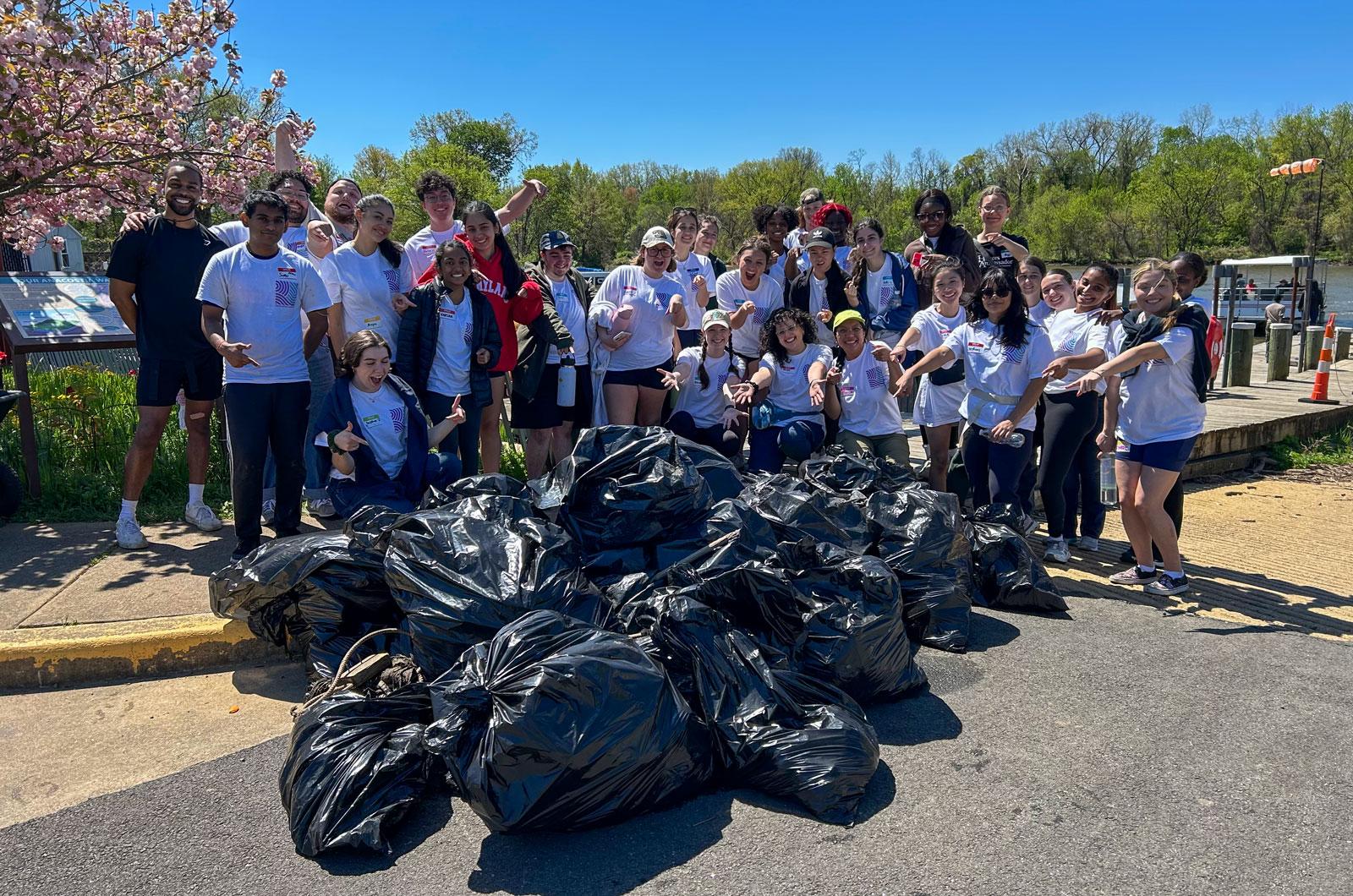 A large group of volunteers poses behind a pile of full trash bags during the Restorative Anacostia River Cleanup cleanup event.