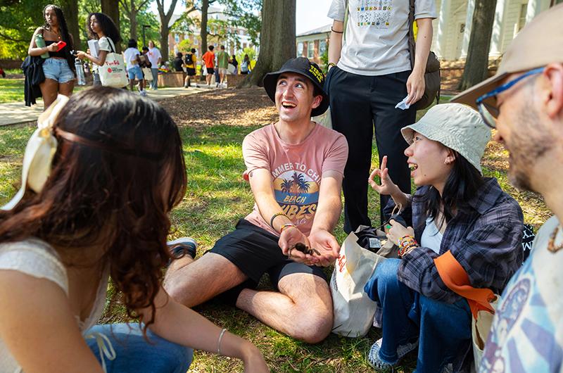 Group of University of Maryland students sitting on the grass and laughing during a sunny outdoor event. One student holds a small animal while others smile and engage in conversation.