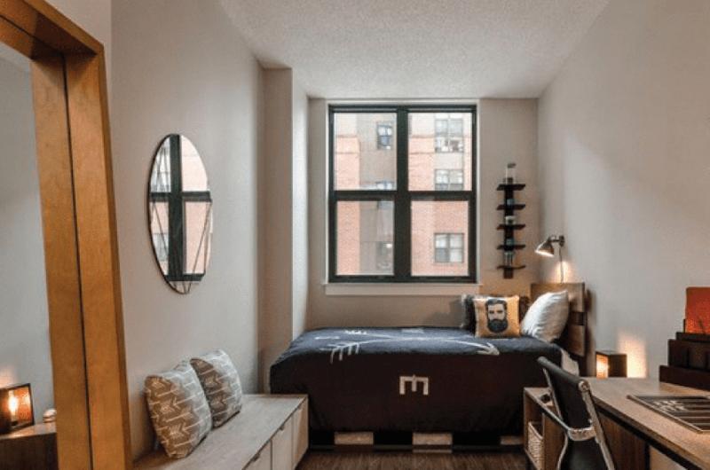 student bedroom with a dark bedspread, circular wall mirror, and large window.