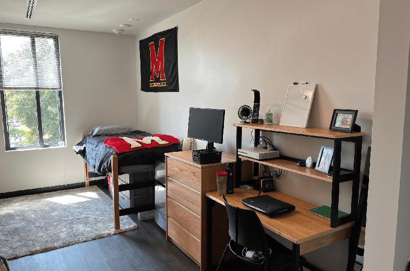 A tidy dorm room with a desk, a "M" flag on the wall, and a bed with a Maryland-themed blanket.