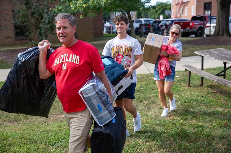 A student and his parents carry luggage and boxes across a campus lawn.