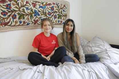 Two smiling roommates sitting together on a bed in front of a colorful floral tapestry.