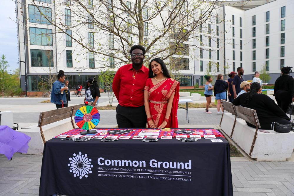 Two students pose behind a "Common Ground" table during the outdoor Heritage Fashion Show event.